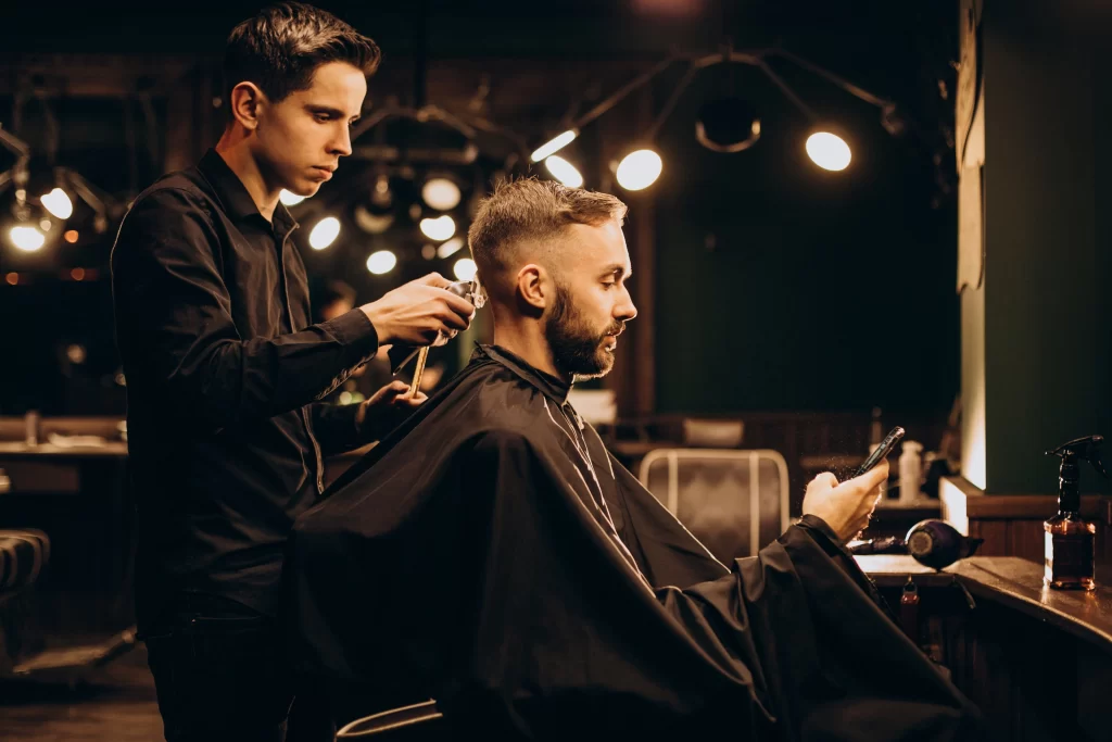 Foto de um barbeiro cortando o cabelo do seu cliente, enquanto ele mexe no celular