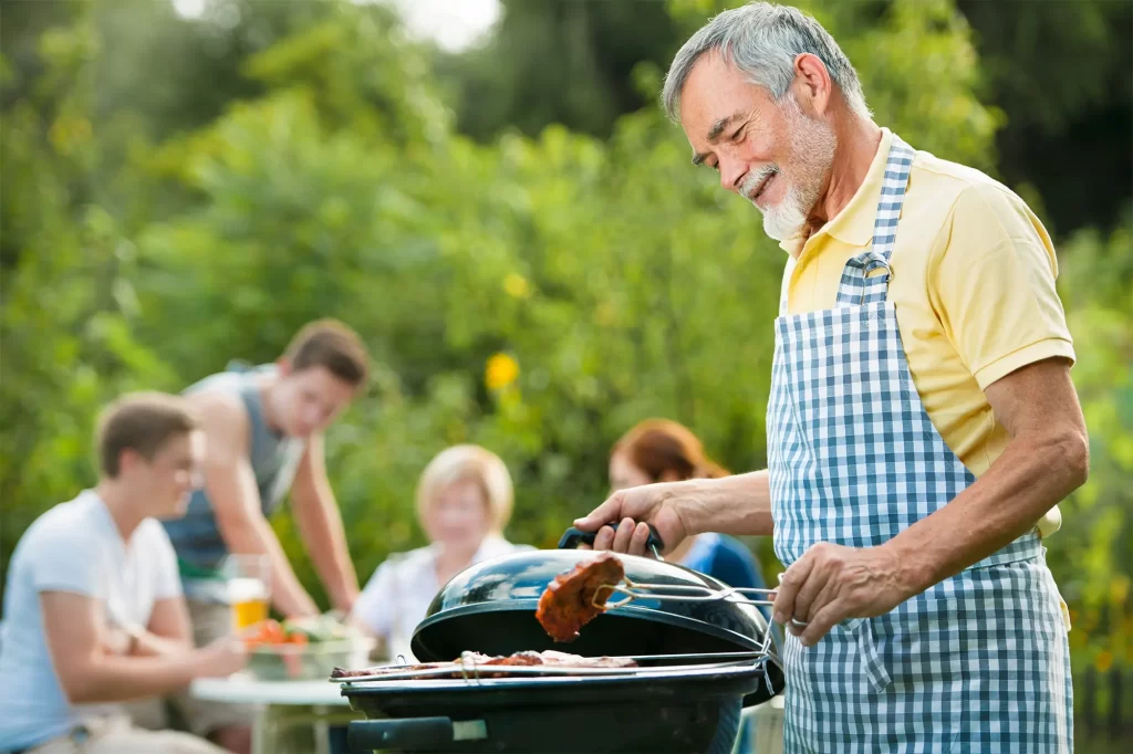 Pai de família fazendo carne em churrasqueira, no fundo, desfocado sua família está sentada em mesa no quintal